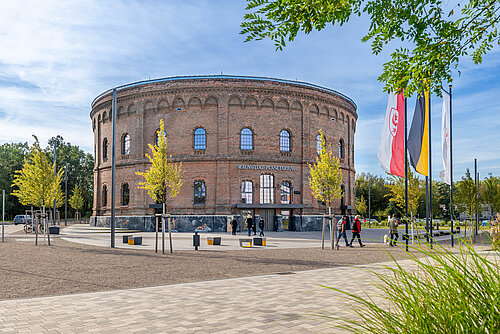 zu sehen ist das ehemaliges Gasometer Halle, ein Zylinderförmiges Backsteingebäude, in dem das Planetarium Halle eingebaut wurde. Im Vordergrund zu sehen Pflanzen, Bänke und Menschen